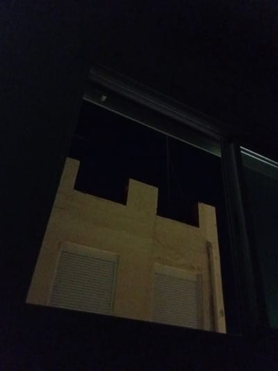 The image shows a view through a window at night in Olula del Río, Spain. The window frame is dark, and the glass reflects a light-colored architectural feature that appears to be a building facade. This facade has a notched or crenellated shape, resembling battlements, with two rectangular window openings that have horizontal blinds or shutters. The rest of the scene outside the window is dark, indicating it is nighttime. The overall impression is a dark, somewhat abstract view of a building at night as seen from inside a room.