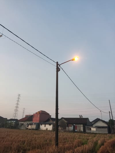 The image depicts a rural or semi-urban scene in Sukorejo, Indonesia, under a pale, hazy sky, possibly at dawn or dusk. A tall, dark lamppost with a lit yellow lamp stands prominently in the foreground, slightly off-center to the right. Several black electrical wires stretch across the frame, attached to the lamppost and disappearing into the distance.

Behind the lamppost, a field of dry, golden-brown stubble, likely harvested rice, stretches towards a row of houses. These houses vary in architectural style and condition. One prominent structure has a distinctive reddish-pink facade with a darker red roof and a small yellow water tank on top. Other houses in the row appear to be a mix of simpler constructions with corrugated metal and brick elements.

In the far left background, a tall, lattice-like electrical pylon is visible, indicating the presence of a power infrastructure within the vicinity. The overall atmosphere is tranquil and suggests a residential area on the outskirts of a populated region. There are no people or animals visible in the frame.