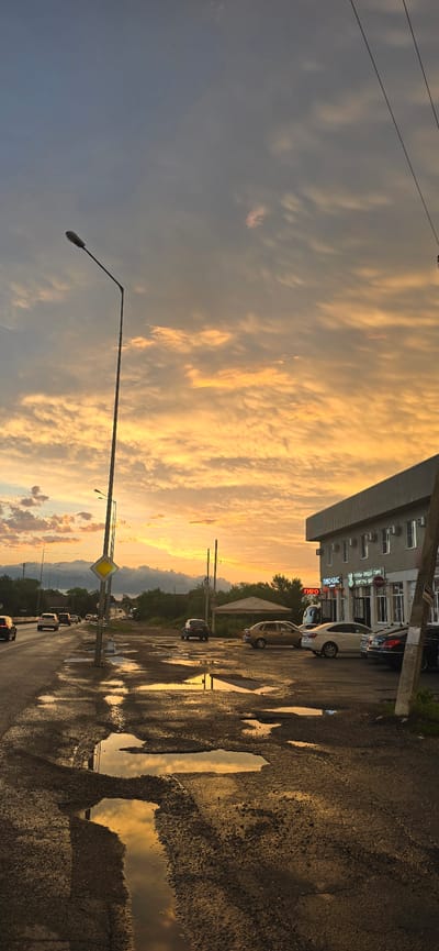 The image depicts a street scene in Suvorovskaya, Russia, during what appears to be sunset. The sky is filled with dramatic, golden-hued clouds, suggesting a warm and vibrant atmosphere. The foreground is dominated by a wet asphalt road, with numerous puddles reflecting the sky and surrounding environment.

Several cars are visible parked along the roadside and driving in the distance, indicating a normal day in the town. A street light stands prominently on the left side of the image, its long arm extending across the frame. In the background, a multi-story building with an awning is present, featuring several windows and air conditioning units. Signs with Russian text are visible on the building, including "ТИРО" and "РОГА-ПИЦЦА-ПИРО". A diamond-shaped yellow road sign with a black border is also positioned near the street. The presence of trees and a glimpse of distant mountains suggest a semi-rural or suburban setting. The overall mood conveyed is one of a peaceful evening, with the dramatic sky adding a touch of natural beauty to the ordinary streetscape.