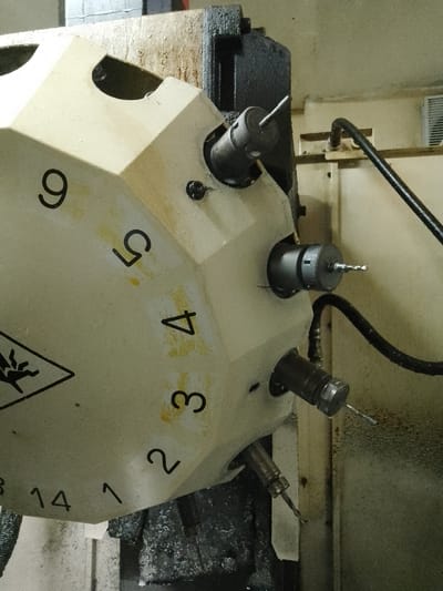 The image displays a close-up view of a multi-spindle rotary tool changer, a component commonly found on CNC machining centers. The tool changer is light beige with black numbers indicating different tool positions, ranging from 1 to 14, arranged in a circular pattern. Several tool holders, each containing a drill bit, are mounted in different positions on the changer. Some of the drill bits are visible, showing their sharpened tips.

The tool changer is attached to a larger, dark grey industrial machine. A black flexible hose, likely for coolant or air, extends from the machine towards the tool changer. The background consists of a wall and some metallic structures, suggesting an industrial or workshop environment. The surfaces of the machine and the surrounding area show signs of use, with some dirt and smudges.

There are no people or animals visible in the image. The scene is static and appears to be an interior shot of a manufacturing facility. The image is an objective depiction of industrial machinery. The context of Ciantra, Indonesia is not directly conveyed by visual elements within the image itself.