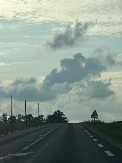 The image captures a view of a road leading uphill towards a horizon obscured by clouds and a hazy sky. Several dark poles with wires stretched between them are visible on the left side of the road, interspersed with dense green trees. On the right side of the road, a triangular warning sign with an exclamation mark is prominently displayed, situated on a grassy embankment. The road itself is paved and marked with white dashed lines indicating lane separation. The sky is filled with a mixture of textured, light-colored clouds and larger, darker, more dramatic cloud formations. There is a faint contrail visible in the upper left quadrant of the sky. No people or vehicles are present in the image. The overall impression is a somewhat overcast day on a rural road in Saint-Maclou-de-Folleville, France.