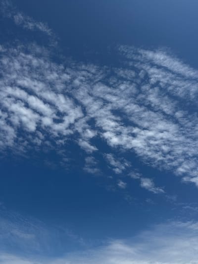 The image shows a bright blue sky with scattered white clouds. The clouds are wispy and thin, appearing in patches across the sky, with some denser formations creating ripple-like patterns. The lighting suggests it is daytime, with the sun likely high in the sky. The scene is devoid of any human presence, structures, or geographical features of Kalngale, Latvia. It is purely a shot of the sky and clouds.