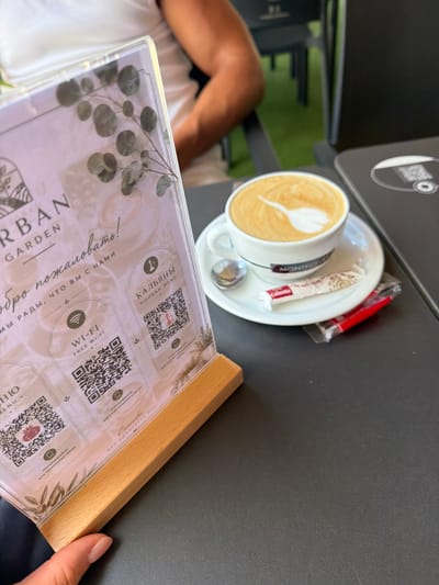 This is a medium shot, taken from a high angle, of a person's hand resting on a menu at a table. The menu is made of clear plastic and is framed by a light-colored wood. The menu displays several QR codes and text in both Russian and English. Visible text includes "URBAN GARDEN," "ДО ПОЖАЛОВАТЬ! МЫ РАДЫ, ЧТО ВЫ С НАМИ" (meaning "Welcome! We are glad that you are with us"), "WI-FI FREE WI-FI," and "КАЛЬЯНЫ HOOKAH MENU."
To the left of the menu, a cup of coffee with latte art is visible, placed on a saucer. A spoon and a sugar packet are also on the saucer. The coffee cup has the word "MONTECELE" printed on it. In the background, a person's arm and leg, wearing a white garment, are visible. The setting appears to be an outdoor cafe, with hints of greenery in the background, possibly a garden area. The overall lighting suggests it is daytime, likely sunny. The scene evokes a relaxed atmosphere, perhaps during a leisurely break. The presence of the menu with QR codes suggests a modern dining establishment. The mention of "La Vila Joiosa, Spain" in the prompt implies the location is a coastal town in Spain.
