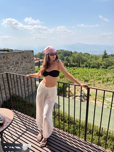 A young woman stands on a balcony overlooking a vineyard. She is wearing a black bikini top, white linen pants, a pink bandana on her head, and sunglasses. The balcony has a metal railing and a table with a small camera. In the background, there are rolling hills covered in vineyards and trees. The sky is blue with scattered clouds. The location is a vineyard in Sassofortino, Italy.