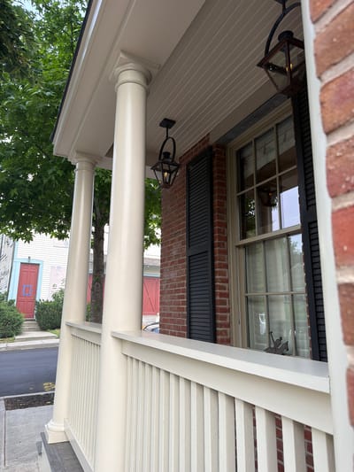 A close-up, ground-level perspective shows a cream-colored porch railing and three fluted Doric columns against a backdrop of a brick building with black shutters and a multi-paned window. A dark metal lantern hangs from the porch ceiling. To the left, a white house with red double doors and light blue trim is visible, with lush green trees and bushes in the foreground. The scene appears to be on a paved street in the Village of Sag Harbor, United States of America, suggesting a residential or historical district.