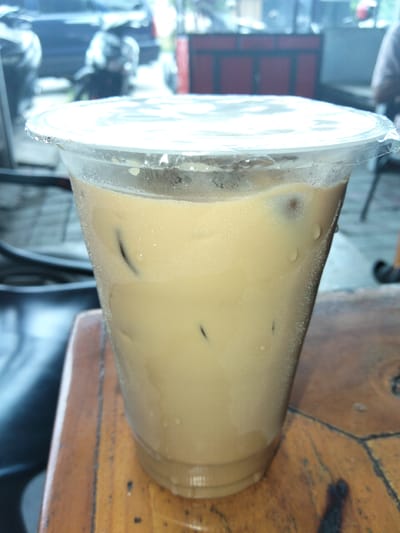 A close-up shot focuses on a clear plastic cup filled with a light brown iced beverage, likely coffee or tea, with ice cubes visible within. The cup is sealed with a clear plastic lid, and condensation has formed on the outside of the cup. The cup rests on a wooden surface, possibly a table, with visible grain and some wear. In the blurred background, elements of an outdoor setting are discernible, including what appears to be a street scene with vehicles and possibly a stall or building. The overall impression is that of a refreshing drink being enjoyed in a casual, possibly commercial, outdoor environment. The location is Geulumpangpayong, Indonesia.