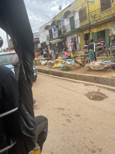The image depicts a street scene in Gwong, Nigeria. In the foreground, a person is seated in what appears to be a tuk-tuk, with part of the vehicle's frame and the person's black clothing visible. The street is paved with asphalt and shows signs of wear, including a prominent pothole filled with muddy water. On the right side of the street, a row of buildings lines the sidewalk. These buildings have multiple stories with balconies, some with metal railings. The facades are painted in various colors, with a prominent yellow building dominating the scene. Shops are open on the ground floor, with green metal shutters. A market is set up along the sidewalk in front of the buildings. Various goods are displayed, including fruits like watermelons and bananas, as well as sacks of produce. Several individuals are present, some appearing to be vendors and others shoppers. A few people are standing or sitting near the market stalls. The sky is overcast.