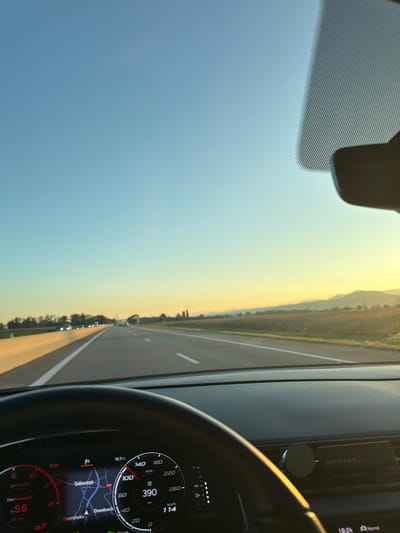 A forensic view from the driver's perspective inside a vehicle travelling on a multi-lane highway in the vicinity of Ebersheim, France, captured during late afternoon or early evening. The dashboard display indicates the vehicle is moving at 100 km/h, has a remaining range of 390 km, and shows an external temperature of 16.5 °C. The time displayed is 18:24. The navigation system explicitly shows "Ebersheim" and "Sélestat" as nearby locations on the route. Visible within the car are the upper portion of a black steering wheel, a digital instrument cluster, and a "RITUALS..." branded air freshener or diffuser attached to the dashboard's right side. Through the windshield, a clear blue sky transitions to a golden-yellow hue at the horizon, where distant hills or mountains are visible. The highway features multiple lanes with white markings. A light-colored concrete barrier runs parallel to the left side of the road, while a large agricultural field occupies the right side, extending towards the horizon. Multiple vehicles are observed ahead on the highway.