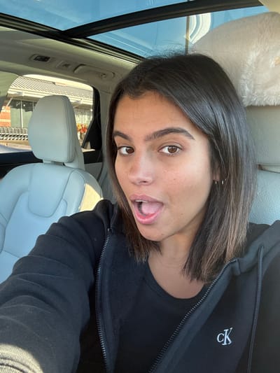 A female individual is depicted in a selfie taken from the front passenger seat of a vehicle, which features light grey leather upholstery and a panoramic glass roof showing a clear blue sky. The subject has dark brown, shoulder-length hair and is wearing a black zippered hoodie with a white "cK" logo on the left chest, over a black top. She looks directly at the camera with an open-mouthed expression, revealing her tongue. A seatbelt is visible across her chest. Through the car window, the exterior of a brick-facade building with a grey roof and an array of shopping carts is visible, indicating a supermarket or large retail store, consistent with a ShopRite location in Lawrence Township, United States. A fluffy, light-colored object is partially visible on the rear passenger headrest.