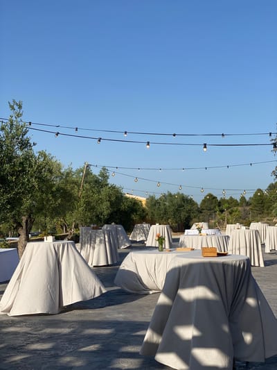 In the city of Bullas, Spain, a paved outdoor area is set up for an event, possibly a wedding reception or outdoor gathering. Numerous round cocktail tables draped with light-colored cloths are arranged across the space. Some tables have small decorative items such as a vase with flowers or wooden boxes. String lights with exposed bulbs are suspended overhead, casting a warm glow in the clear blue sky. The background features lush green trees, including olive trees, and a hint of foliage further in the distance. The overall atmosphere is serene and organized, suggesting preparations for a daytime event.