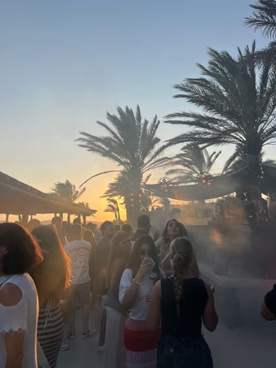 The image depicts a crowded outdoor scene at sunset in Psarou, Greece, likely within a beach club or resort environment. Numerous individuals, predominantly young adults, are gathered, standing and mingling. Several tall palm trees are silhouetted against the sky, which transitions from light blue at the top to a vibrant orange and yellow hue near the horizon where the sun is setting.
In the foreground and midground, a dense crowd is visible. A female subject in a white t-shirt and red patterned skirt is centrally located, observed drinking from a wine glass. To her right, another female subject with blonde hair makes a distinct "kiss" expression toward the camera. To her left, a male subject is visible from behind wearing a white t-shirt with "EAGLE POINT" printed on the back. Mist or smoke emanates from the lower right portion of the scene, partially obscuring the base of a large, elevated structure that appears to be a stage or DJ booth equipped with large speakers. To the far left, a section of a covered, open-air structure with a flat roof is visible. The ground appears to be a solid, paved surface.