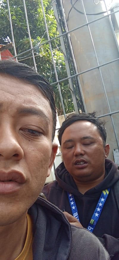 Two men are captured in a close-up, selfie-style photograph outdoors, with a chain-link fence prominently in the foreground. The man on the left is closer to the camera, his face partially obscured by the edge of the frame. He appears to be looking towards the camera, with his mouth slightly parted and his eyes open. The man on the right is positioned behind and to the right of the first man, his eyes closed, and his mouth open as if speaking or reacting. He is wearing a dark hoodie with a lanyard featuring stars and a blue, yellow, and green emblem. In the background, lush green foliage and a concrete structure are visible through the fence. The overall lighting suggests it is daytime, and the setting appears to be an urban environment in Kiangroke, Indonesia, likely near a building or enclosed area due to the fence.