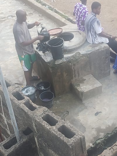In Alalubosa, Nigeria, a high-angle shot depicts a concrete structure, likely a well, where multiple black buckets are positioned. One man is actively drawing water from the well using a bucket, while two other men are standing nearby, observing. The surrounding environment appears to be a residential area with basic infrastructure.