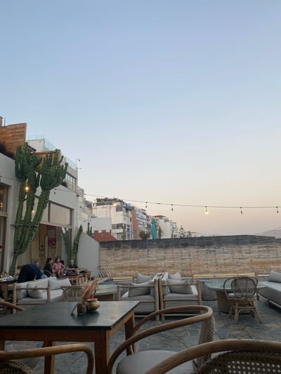 The image depicts an outdoor seating area at a restaurant or lounge in Taghazout, Morocco. The setting appears to be on a rooftop or elevated terrace, overlooking a cityscape of buildings with a clear, pale blue sky above. The architecture visible in the background is a mix of modern and traditional styles, with white-washed walls and balconies.
In the foreground, there are several seating arrangements with light-colored sofas and chairs, made of rattan and wood. A dark-topped table in the immediate foreground holds a small vase with dried flowers and reeds. To the left, a large cactus is prominently featured against the building.
A group of people are seated on a sofa in the mid-ground, engaged in conversation. The overall atmosphere suggests a relaxed evening gathering, possibly during sunset, indicated by the soft lighting and the colors of the sky. String lights are visible along a wire stretched across the scene, contributing to the ambiance. A swimming pool is partially visible to the right, suggesting a resort or hotel setting. The ground is paved with stone tiles.