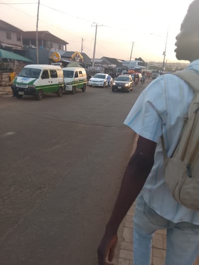 This image captures a vibrant street scene in Oke Ogun, Nigeria, during what appears to be the late afternoon or early evening, characterized by a soft, warm, and somewhat hazy light from a low sun.
In the foreground, a person is visible from the right side, partially obscuring the view. They have dark skin, are wearing a light blue and white striped short-sleeved shirt, light-colored jeans, and a tan backpack slung over their right shoulder. Their head is turned to the left, and their right arm is visible.
The mid-ground features a bustling road with several vehicles. Two prominent white and green minibuses are parked or moving slowly on the left side of the road. These vehicles are commonly used for public transport in Nigeria. Further down the road, a black sedan with its headlights on, a white sedan, and what appear to be several motorcycles are also visible, indicating ongoing traffic. The road itself is paved but shows signs of wear.
Along the left side of the road, several buildings are visible, some two-storied, with various stalls and awnings in front of them, suggesting commercial activity. One circular sign clearly displays the word "SHAWARMA," indicating a food vendor. Another sign in the distance has "ASSUNNAH" written on it. Utility poles and overhead wires crisscross the sky. The overall atmosphere suggests a typical, active urban or semi-urban environment. No specific emotions are discernible; it's a candid snapshot of daily life.