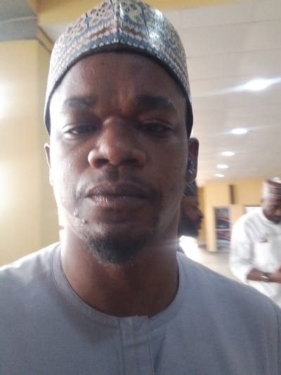 This image captures a close-up portrait, likely a selfie, of an adult male in an indoor setting within Kwi, Nigeria. The male subject, positioned centrally, has dark skin, a short beard, and appears to have two small blemishes or piercings on his left chin area. He wears a light grey traditional garment with a stand collar and a patterned cap (kufi-style) featuring blue, brown, and cream geometric designs. His gaze is directed towards the camera. The background is out of focus, revealing a light-colored ceiling with recessed lighting and light-colored walls. Partially visible in the blurred background on the right is a second male figure in a white traditional garment and a similar cap, seemingly looking downwards. A third, less distinct figure is also faintly visible in the mid-ground.