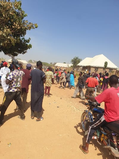 A large crowd congregates near a building with a white roof. People are dressed in casual attire, their figures small against the landscape. A man wearing red sits on a motorbike in the foreground, his sandal-clad foot visible. The ground appears dry and dusty. Trees stand on the left side of the image, adding some greenery. The sky is clear and blue.