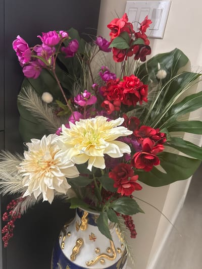 An artificial floral arrangement is displayed in a decorative vase. The arrangement features a variety of flowers including large cream-colored dahlias, red roses, and clusters of smaller pink and red blossoms. Green monstera leaves and wispy, feathery stalks are also incorporated. Red berries are interspersed throughout the bouquet. The vase is white with blue and gold detailing. The background consists of a dark surface on the left and a light-colored wall with a light switch on the right. The image is an indoor still life, and there is no indication of the specific location in Easton, United States of America.