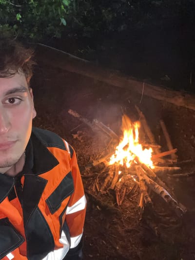 The image depicts a person lying down next to a bonfire. The individual is wearing an orange and black jacket with reflective stripes. The bonfire is built with logs and is actively burning, casting light on the surrounding area. The background is dark and suggests a wooded area or a campsite. The photograph appears to be taken at night or in low light conditions. There is no definitive evidence from the image that specifies the location is Krasnyi Jar, Russia.