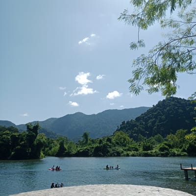 A wide shot captures a serene natural landscape with a body of water in the foreground and lush, tree-covered mountains in the background under a clear, bright blue sky. Several small boats, including canoes or kayaks with people paddling, are scattered across the calm water. In the lower foreground, a stone pathway leads towards the water. A wooden dock with two vertical posts extends into the water on the right side of the frame. The overall scene suggests an outdoor recreational area, possibly a lake or a calm river in a rural or natural setting.