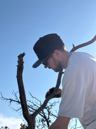 The image captures a male individual outdoors in the Unincorporated Santa Monica Mountains, United States of America. The subject is presented in profile, facing slightly downwards and towards the left of the frame. He wears a dark baseball cap, dark sunglasses, and a light-colored (likely white) short-sleeved t-shirt, and has facial hair. His right hand is visible, grasping a long, bare tree branch. To the left of the subject, a prominent, gnarled, dry tree trunk or thick branch stands vertically. The background consists of a clear, bright blue sky with sparse, bare tree branches visible against it.