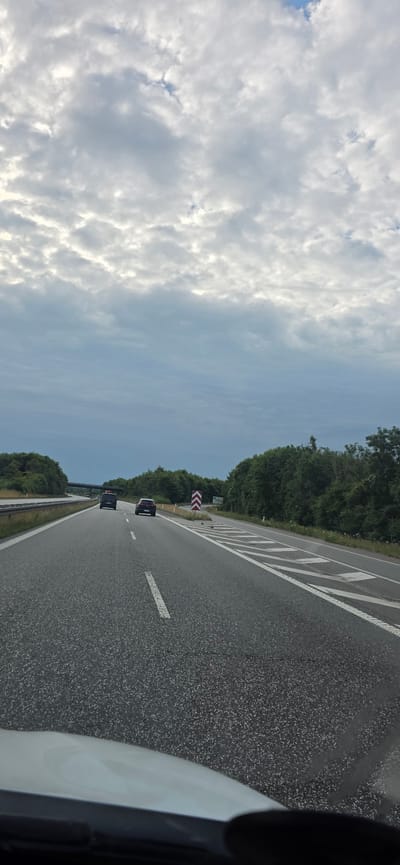 The image shows a perspective from inside a vehicle traveling on a highway in Vejen, Denmark. The highway has multiple lanes and is bordered by dense green trees on both sides. The sky is overcast with a textured pattern of clouds. Two vehicles are visible on the highway ahead, a black car and a dark colored van or SUV. A red and white chevron sign is placed on the right side of the road. The road surface is asphalt and lane markings are visible. The front hood of the vehicle from which the photo is taken is visible in the foreground.
