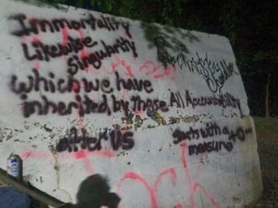 A nighttime photograph captures a white concrete wall heavily adorned with graffiti. The primary text, written in black spray paint, includes phrases such as "Immortality," "Likewise Singularity," "Which we have inherited by These.," "after us," and "All Accountability Starts with a zero-measure." Red spray paint is also visible, used for drips, underlines, and additional, less legible tags and symbols, including what appears to be a heart shape. Overlapping and fainter black graffiti is present, with some parts resembling a tag like "Slank Daddy Chandler." A light-colored spray paint can rests on the ground at the bottom left of the wall, suggesting recent graffiti activity. The background is dark, indicating a nighttime setting, and some vegetation is faintly visible beyond the wall. A dark shadow, possibly from a person, is cast onto the bottom of the wall in the foreground. The scene depicts an outdoor environment, likely in Tahlequah, United States of America.