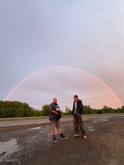 This media file shows a wide shot of a natural landscape featuring two men and a rainbow. The setting appears to be an outdoor location with a gravelly ground and trees in the background. The sky is overcast with a faint rainbow arching across it, suggesting that it has recently rained or is about to rain.
In the foreground, two men are standing on the gravel. The man closer to the bottom of the frame is wearing a plaid shirt and shorts, with a cigarette in his hand. The man above him is wearing a jacket over a t-shirt and trousers. Both men are looking towards the camera.
The lighting in the image is soft and diffused, typical of an overcast day. The time of day is likely late afternoon or early evening, given the soft light and the presence of the rainbow. There are puddles on the ground, indicating recent rainfall. The overall mood of the image is calm and serene, enhanced by the natural beauty of the rainbow.
There is no visible text in the image.