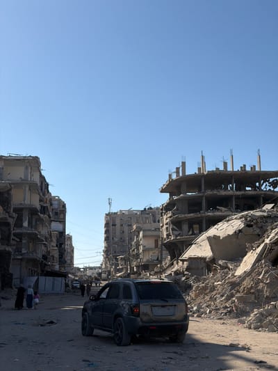 The image shows a street in the city of Jabalia, Palestinian Territories, lined with severely damaged buildings. A dark-colored SUV is parked on the right side of the dirt road, facing away from the camera. In the background, several multi-story buildings are in ruins, with exposed concrete structures and debris. Several people, some wearing traditional clothing, are visible walking on the road and near the damaged buildings. The sky is clear and blue. The scene depicts widespread destruction, consistent with an area experiencing conflict or significant damage.