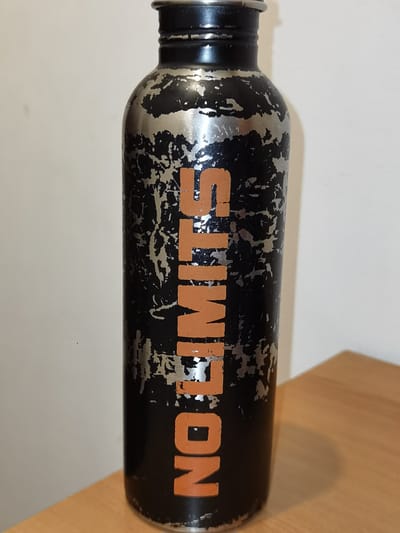 A black water bottle with the words "NO LIMITS" written vertically in orange is shown against a plain wall. The bottle has a distressed metallic silver pattern overlaid on the black surface, suggesting wear and tear. The bottle is standing upright on a wooden surface, which appears to be a table or desk. The lighting is soft, casting a slight shadow behind the bottle. There are no people present in the image. The location is not specified in Veria Municipality, Greece.