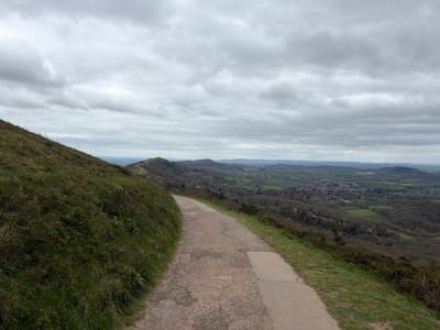 The image depicts a rural scene in the Malvern Hills, United Kingdom, under a cloudy sky. A gravel and concrete pathway stretches into the distance, winding along a hillside covered with grass and sparse vegetation on the left. The path offers a wide-angle view of a landscape comprised of rolling hills, wooded areas, and fields. A town or village is visible in the distance. No people or animals are visible in the foreground or midground.