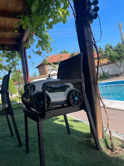 A white and black robotic lawnmower, identified by the "WYBOT" logo, sits on a black plastic chair under a wooden pergola. The chair and lawnmower are positioned in a yard with artificial grass, adjacent to a swimming pool. In the background, a building with a red tile roof and trees are visible. The scene appears to be outdoors in daylight, with a clear blue sky. This location is in Totleben, Bulgaria.