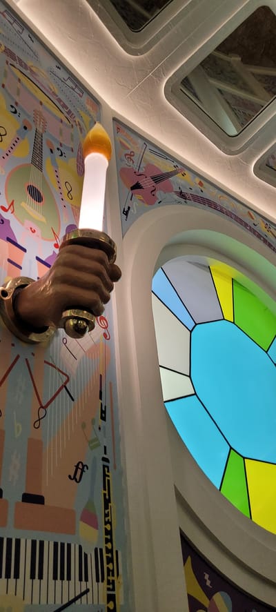 This image shows a close-up of a wall decoration and a stained-glass window in Wattens, Austria, likely within the Swarovski Crystal Worlds. On the left, a bronze-colored arm holds a light fixture that resembles a torch with a translucent white flame that is illuminated. The arm is mounted on a wall adorned with a whimsical, colorful mural depicting musical instruments and notes, including a guitar, a violin, piano keys, and musical symbols like treble clefs. The mural has a light blue background with abstract shapes and lines in various colors.
To the right, a portion of a large, circular stained-glass window is visible. The window is divided into geometric sections of vibrant colors, including bright blue, yellow, green, and off-white, outlined by thick black lines. The window appears to be set within a curved architectural element. The ceiling above shows recessed lighting and mirrored panels, reflecting the decor. The overall impression is one of artistic and playful design, characteristic of the Swarovski Crystal Worlds.