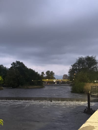 The image shows a river flowing in the city of Komgaron, Russia. The view includes a cityscape faintly visible in the distance, framed by dark clouds. There are lights along the riverbank. Vegetation lines both sides of the river. The water flows from a higher level to a lower level. There are no people visible in the image.