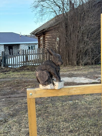 The image depicts a rural scene in Дулесово, Russia. A carved wooden rabbit sculpture rests on a wooden block, which is positioned on a wooden frame. In the background, there's a rustic wooden building with bare branches obscuring parts of it. Adjacent to the wooden building is a brick structure with a grey roof and a wooden fence in front. The ground is covered in dry grass and dirt.