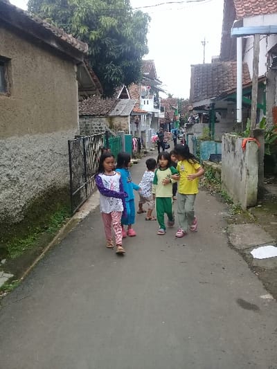 The image shows a narrow alleyway in the city of Kecamatan Cimaung, Indonesia, located at latitude: -7.07, longitude: 107.555. Six children are walking in a single file line on a gray, paved surface. The children are all wearing casual clothing and appear to be young. The alleyway is lined with houses and other buildings on either side. There is lush greenery in the background. The children are walking toward the camera. The children are not interacting with each other. The sky is overcast. There is no indication of time of day.
