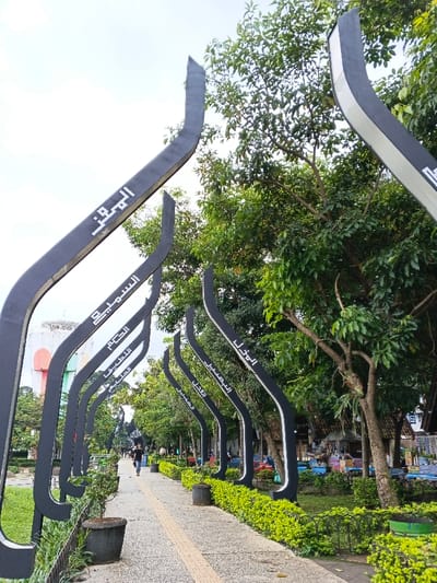 A pathway lined with black, curved metal structures featuring Arabic text runs through a park. Lush greenery borders the path, with trees overhead creating partial shade. People stroll along the walkway, and a building with a colorful facade is visible in the distance. Potted plants and manicured bushes decorate the edges of the path, adding to the park's inviting ambiance.