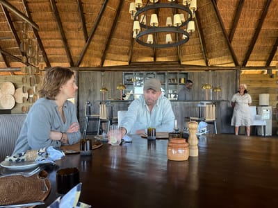 The image shows two people seated at a large wooden table in what appears to be a restaurant or dining area in Khwai, Botswana. The woman on the left is wearing a gray shirt and has her arms crossed. The man on the right is wearing a light-colored shirt and a baseball cap, and he's holding a glass of liquid with a lemon slice. The table is set for dining with plates, silverware, woven placemats, and napkins. A salt and pepper grinder and a ceramic jar are also on the table. Behind them, there's a bar area with two men in uniform working. A woman in uniform stands near a chair and some storage cabinets. The building has a thatched roof and wooden walls. A chandelier hangs from the roof above the table. Decorative woven items are visible on the wall to the left.