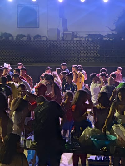 The image depicts a crowded outdoor gathering at night in Blanca, Spain, illuminated by bright blue lights from above. A diverse group of individuals, primarily young adults, are seen mingling and standing in close proximity. Many are dressed in casual attire, including t-shirts, shorts, and athletic wear. Some individuals are holding drinks in plastic cups. In the foreground, a table is laden with various items, including plastic bags, bottles of water, a teal bowl, and what appears to be a cooler bag underneath. The background features a building with a distinctive arched window structure, and a latticed fence. The overall atmosphere suggests a festive or social event.