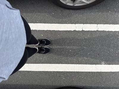 An overhead image depicts an individual standing on an asphalt surface, likely a parking lot, in Plainsboro Township, United States. The subject is wearing a grey top, black leggings, and black athletic shoes with visible white soles. Two parallel, worn white painted lines traverse the asphalt horizontally, positioning the individual between them. A partial silver multi-spoke vehicle wheel is visible in the upper right quadrant, and a section of a black vehicle tire is present in the lower left, indicating the presence of adjacent parked vehicles. The scene is illuminated by natural outdoor light.
