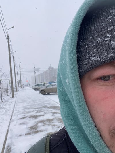 A low-angle, rotated image shows a partial view of a person's face on the upper side, revealing their right eye, brow, forehead, and the top of their head covered with a gray knit cap. The person is wearing a light green hooded garment, likely a jacket, with snow clinging to the fabric. Below the person, the image captures a snow-covered city street. Cars are parked along the right side of the street, and a building with a distinctive architectural style can be seen in the background, partially obscured by snow. Utility poles and wires run along the left side of the street, further suggesting an urban setting. The overall impression is of a snowy day in a city environment. The location is Chemoshur, Russia.