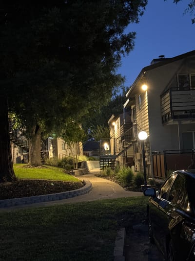 In the city of Antelope, United States, at dusk, a residential complex with two-story buildings is depicted. The buildings feature wood-paneled siding and balconies with metal railings. Exterior staircases lead to the upper floors. Streetlights, one with a spherical globe, illuminate the pathways and the surrounding grass. A large tree with dense foliage stands to the left, casting shadows. A black car is parked on the right side of the frame. The sky is a deep twilight blue.