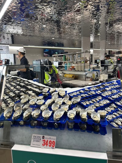 Rows of fresh oysters displayed in blue plastic trays on a bed of ice, a price tag indicates "$34.99" for "Pacific Oyster Jumbo." An employee wearing a white hat and a dark uniform is facing the oysters. Another employee wearing a yellow vest and pink gloves appears to be restocking or preparing the oyster display. The setting is likely a seafood market or grocery store. The ceiling is mirrored.