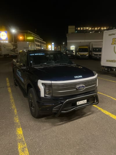 A dark blue Ford F-150 Lightning electric pickup truck is centrally parked at night in an asphalt parking lot with yellow markings in Volketswil, Switzerland. The truck features a black front bull bar and displays the Swiss license plate "ZH 31294 U" on its bumper. The word "FRUNK" is illuminated in white text on the vehicle's hood, and "F-150 LIGHTNING" is visible in light blue text on its side. A white sticker displaying "FR. 88'900.-" is affixed to the upper part of the windshield. The truck's front lighting signature, including a full-width white light strip, is active. In the background to the left, a commercial building with illuminated "Lidl" and "Goodyear" signs is visible, alongside multiple parked white delivery trucks. To the right, a yellow van, a white van, and a large white delivery truck labeled "HOME2H" are parked, with a multi-story building featuring illuminated windows on its upper floors situated behind them. No individuals are present.