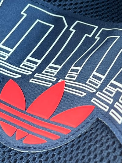 A close-up, high-angle shot shows a portion of a navy blue fabric item, likely a bag or clothing, featuring white outlined lettering and a red Adidas logo. The lettering appears to spell out "TEAM" with each letter formed by stacked rectangular shapes. The Adidas logo, a trefoil, is prominently displayed in bright red. The background is a dark, textured mesh fabric. The image does not provide any information about the city of Kinawley or the United Kingdom.