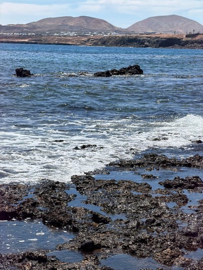 This horizontally oriented image captures a rugged coastal landscape in Haría, Spain, under clear, sunny skies. The foreground features an intricate arrangement of dark, porous volcanic rock formations, characteristic of lava flows, which are partially submerged in the ocean. These rocks are wet and glisten with sunlight, displaying their textured surfaces. White, foamy waves crash against or recede from the rock formations, while calmer sections of water reflect the bright light.

The midground consists of the open ocean with gentle ripples and shimmering reflections from the sun. A few isolated, dark rock outcrops are visible within the water further from the shore.

In the background, a dry, mountainous coastline rises under a light blue sky dotted with scattered white clouds. The mountains appear barren and brownish-grey. Along their base, a cluster of white buildings indicates a small town or settlement, nestled between the arid land and the sea. The image depicts a bright, peaceful daytime scene, emphasizing the natural beauty and geological features of the volcanic coast. No human activity or visible text is present.