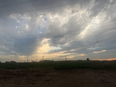 This image captures a cloudy sky over a rural landscape in Boralday, Kazakhstan. The sky is filled with dramatic, textured clouds, with patches of sunlight breaking through, suggesting either dawn or dusk. Several power lines crisscross the frame, indicating the presence of electrical infrastructure in the area.
In the middle ground, a band of dark green vegetation, likely fields or crops, stretches across the horizon. Beyond the vegetation, a line of utility poles stands silhouetted against the sky, further emphasizing the rural and possibly agricultural nature of the setting. The foreground is dominated by a mound of dark brown earth, possibly a construction site or freshly tilled soil, adding a rugged texture to the scene.
The overall mood of the image is somewhat dramatic and tranquil, with the interplay of light and shadow in the clouds creating a sense of depth and atmosphere. There are no people or identifiable animals visible in the frame. The time of day is likely either early morning or late evening, given the quality of the light and the cloud cover. No text is visible in the image.