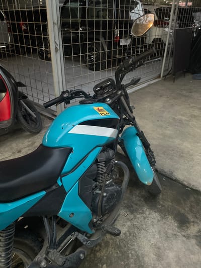 A teal-colored Honda motorcycle, featuring a black seat and handlebars, is parked on a concrete surface in Lhok Mereubo, Indonesia. The motorcycle's fuel tank is adorned with a white and black stripe decal, and a yellow sticker with "PLN" in black lettering is affixed to the upper right side of the tank. The front mudguard is also teal. To the left, a portion of a red and black scooter is visible. Behind the motorcycles, a gray metal grid fence separates the foreground from a background containing multiple dark-colored vehicles, including a truck or SUV with a license plate reading "BL 1996 ZZ". The concrete ground is weathered and exhibits some staining. No individuals are present in the scene.