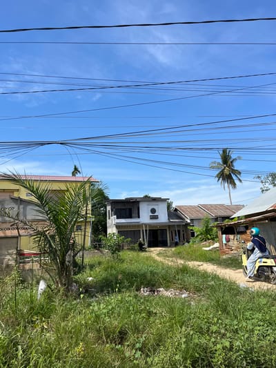The image shows a scene in Dalam, Indonesia, with houses, a dirt road, and lush greenery under a clear blue sky. The perspective is from ground level, looking up and slightly to the side.

In the foreground and left side of the image, a dense network of electrical wires stretches across the sky. To the right of the wires, several houses are visible, with varying architectural styles. One house is white with dark windows and a circular detail above the door, labeled "CP STUDIO." Another house is yellow with a reddish roof. Further back, a tall coconut palm tree stands out against the blue sky.

On the dirt road, a yellow motorbike is parked, and a person wearing a blue helmet and a blue shirt with white patterns is sitting on it, possibly taking a break or preparing to leave. A piece of blue fabric is draped over their lap. To the left of the motorbike, a small, rustic structure made of corrugated metal is present, with some laundry hanging on it.

The scene appears to be during the daytime, with bright sunlight casting shadows and highlighting the vibrant green of the grass and vegetation. The weather seems fair. The overall impression is one of a residential area in a tropical or subtropical region, characterized by a mix of modern and simpler structures, abundant plant life, and the presence of utility infrastructure. The presence of trash scattered along the edge of the road indicates potential environmental concerns in the area.