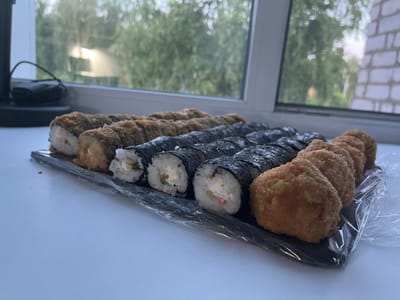 A tray of sushi rolls sits on a white windowsill, with a window and trees visible in the background. The sushi is arranged in two rows. The front row consists of several fried sushi rolls, while the back row contains several maki rolls. The tray is covered in plastic wrap. The image appears to have been taken indoors, possibly in a residential setting in Yaransk, Russia, given the presence of a brick wall visible through the window.